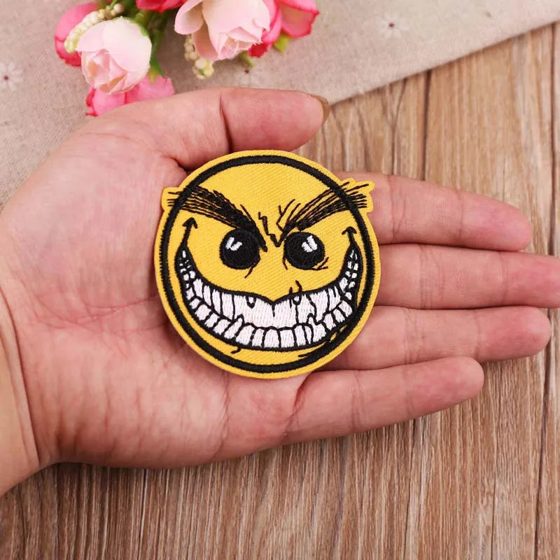 A hand holds a yellow embroidered patch featuring a menacing, grinning face with sharp teeth and angry eyes.