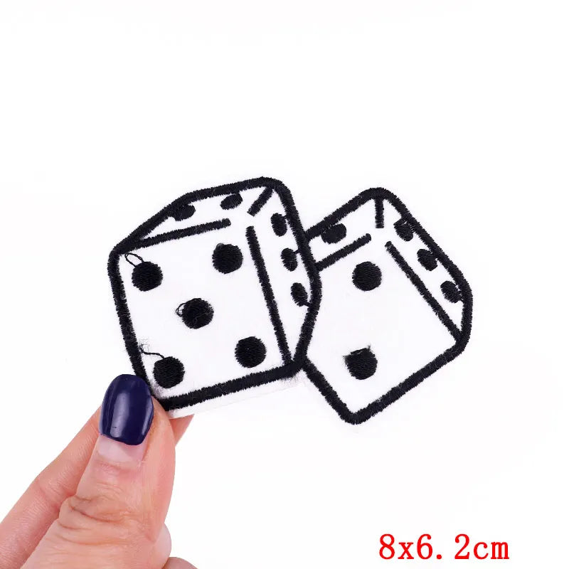 A hand holds two embroidered black and white polka dot dice patches, measuring 8x6.2 cm.