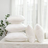 A stack of white cotton-filled decorative throw pillow inserts resting on a white surface near a window.