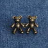 A pair of antique bronze bear button pins, one holding the other, resting on a blue denim fabric.