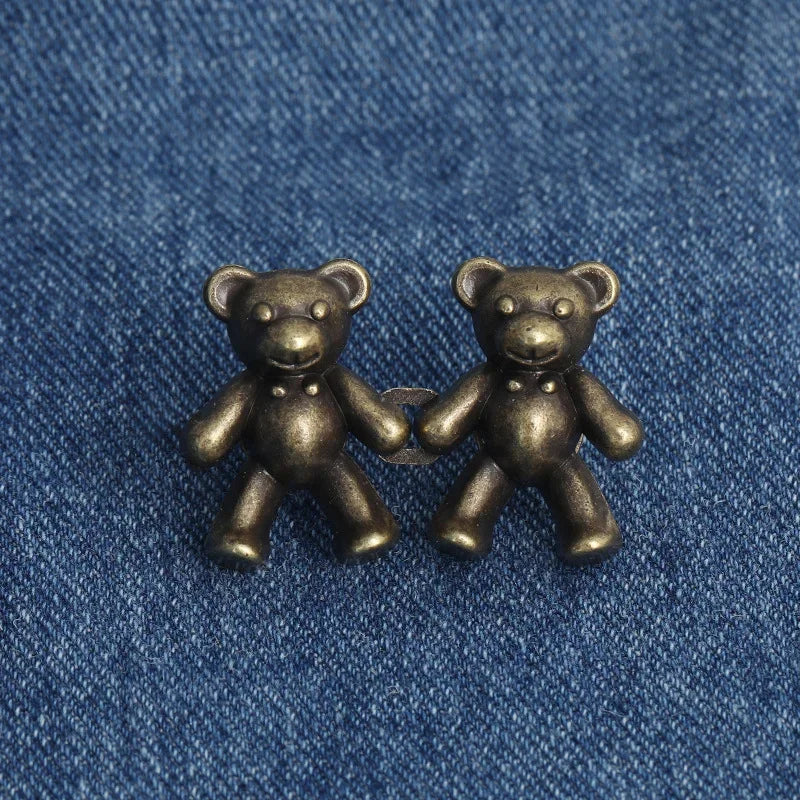 A pair of antique bronze bear button pins, one holding the other, resting on a blue denim fabric.