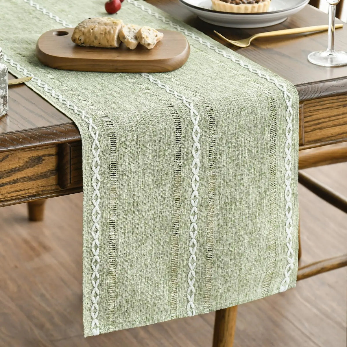 A light green embroidered cotton linen table runner with decorative tassels placed on a wooden table.