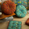 A vibrant, round bohemian cotton linen throw cushion in orange and teal patterns sits on a wooden shelf.