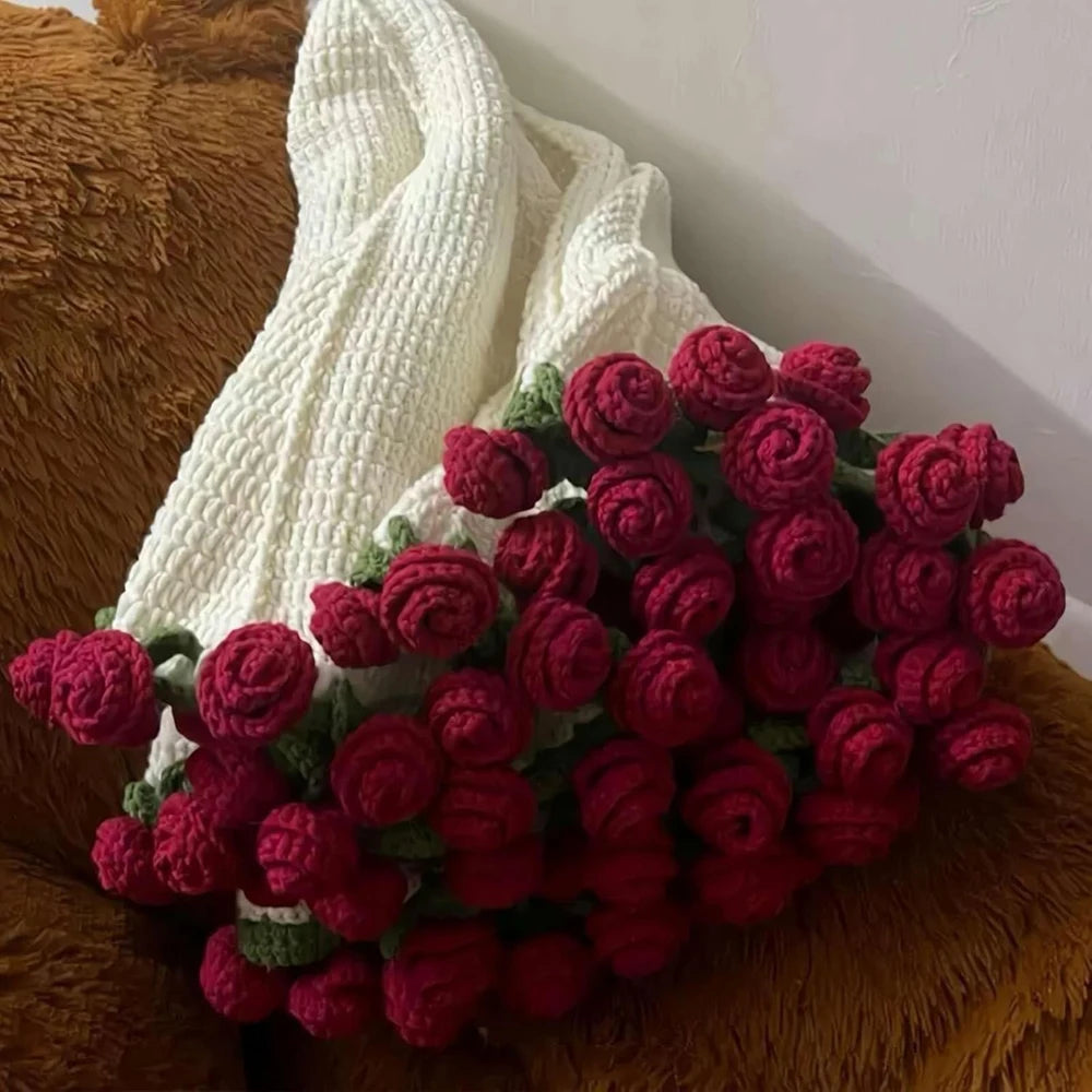 A bouquet of handmade rose crochet flowers in shades of red and green displayed next to a white knitted blanket.