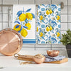 A set of two blue-edged lemon kitchen towels featuring yellow lemons and blue foliage, displayed next to a copper plate and b
