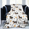 A light-colored flannel blanket featuring a repeating pattern of brown dachshund silhouettes and paw prints draped over a dar