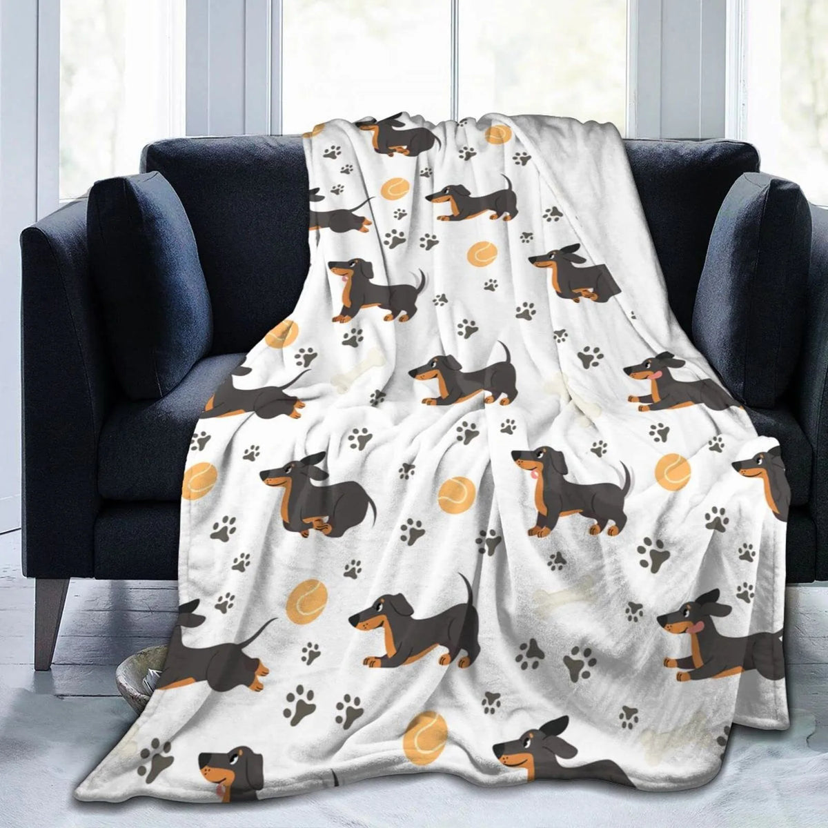 A light-colored flannel blanket featuring a repeating pattern of brown dachshund silhouettes and paw prints draped over a dar