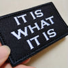 Black embroidered patch with white text reading IT IS WHAT IT IS
