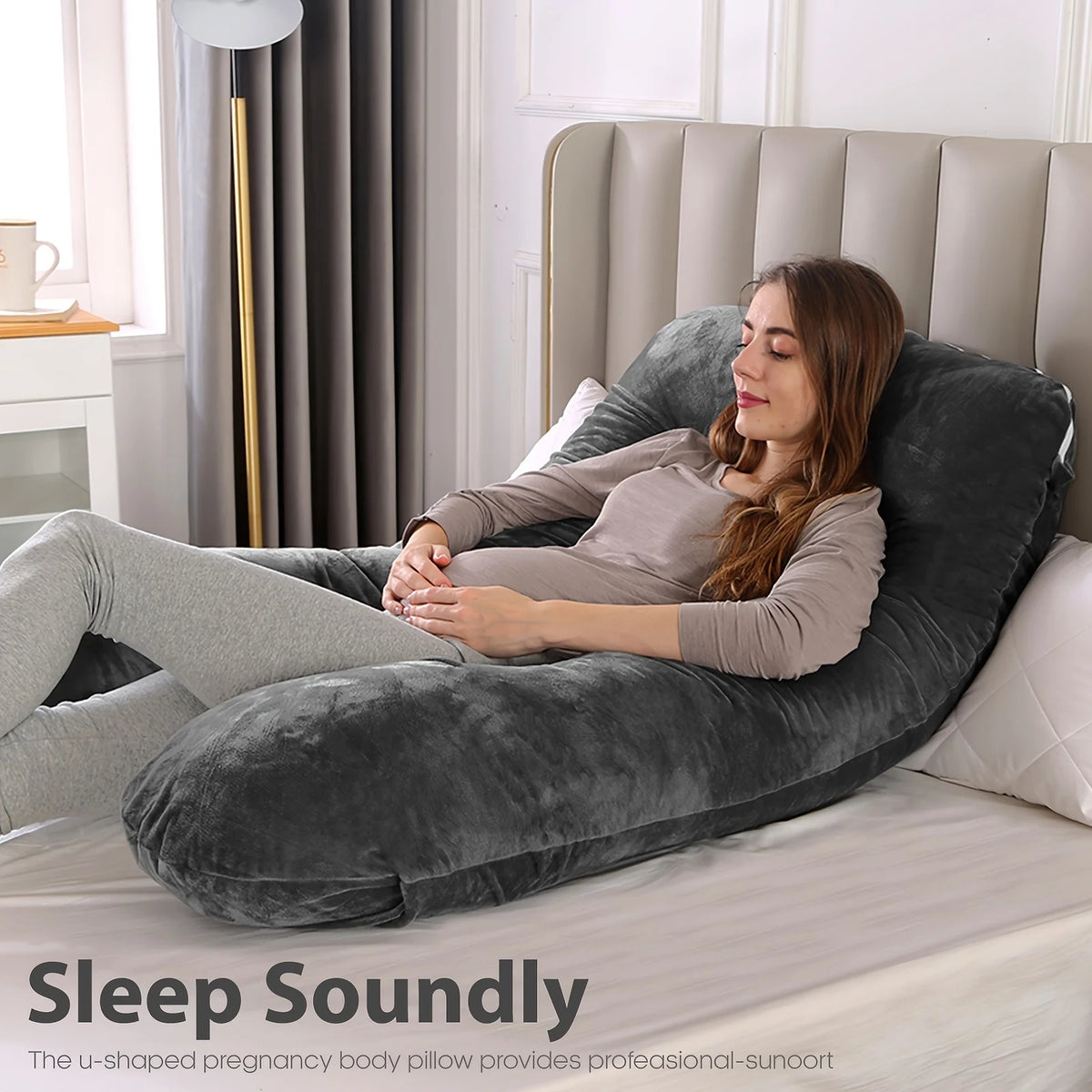 A woman rests her head on a gray U-shaped pregnancy body pillow on a bed.