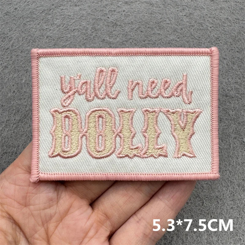 Pink embroidered patch with the text All need Dolly on a light pink fabric background