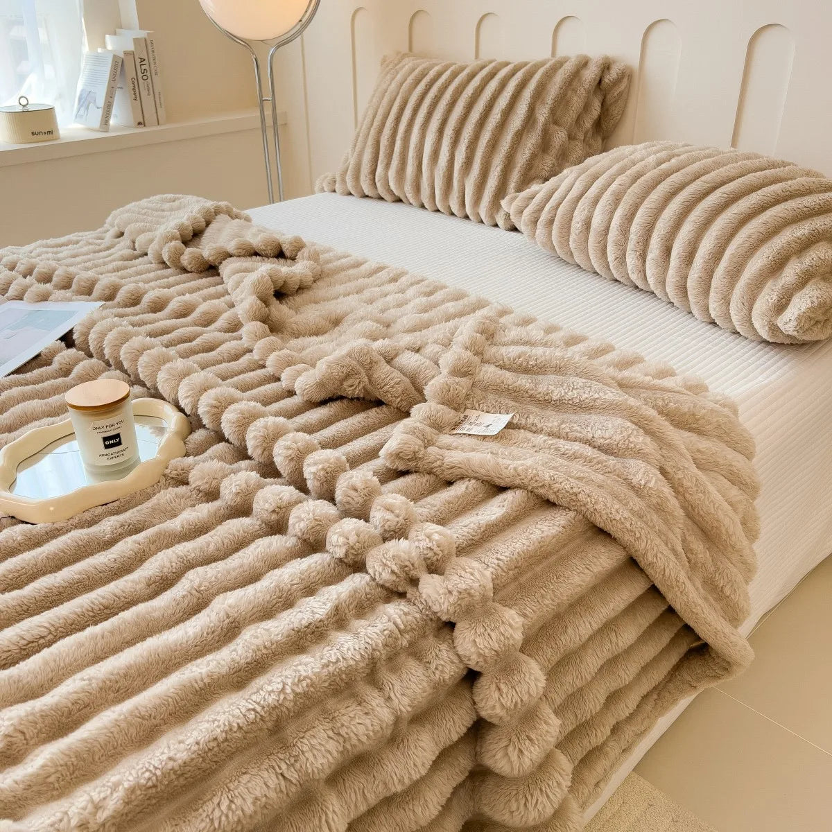A thick, beige, fuzzy blanket is draped over a white bed with matching knitted pillows.