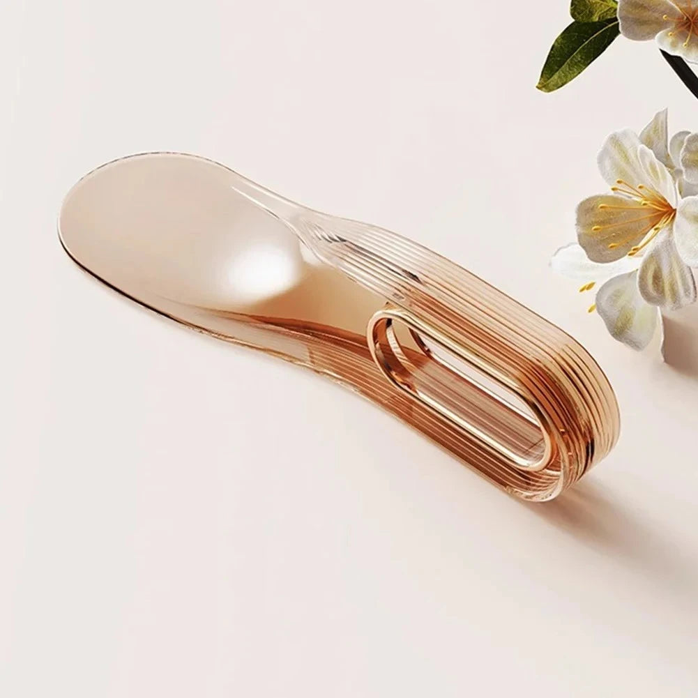 A rose gold mattress lifter wedge is shown on a light surface next to some green leaves and white flowers.