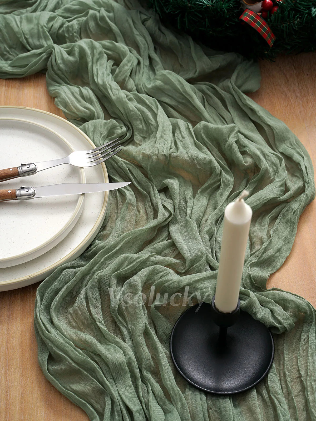A green crinkled cheesecloth table runner draped over a wooden surface with cutlery and a white candle.