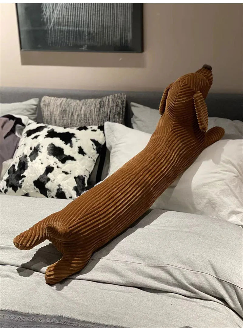 A long, brown corduroy dachshund body pillow resting on a white bed with pillows.