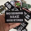 A hand holds a black iron-on slogan patch that reads BAD DECISIONS MAKE GOOD STORIES
