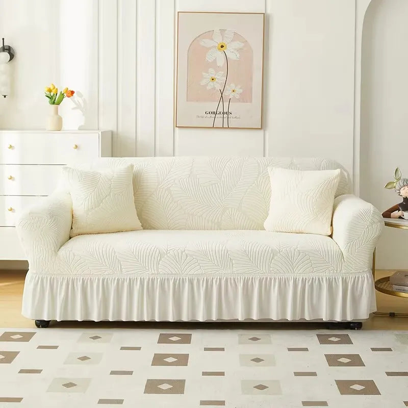 A cream-colored jacquard elastic sofa cover with a gathered skirt displayed in a bright, modern living room.