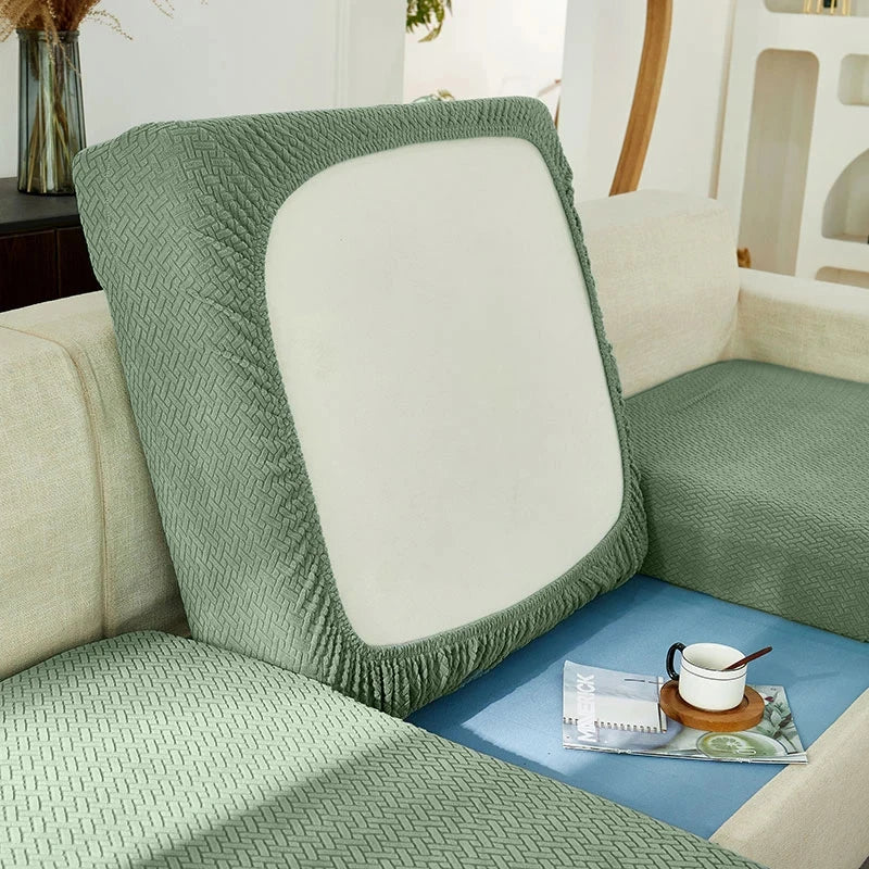 A close-up of a sage green jacquard sofa slipcover with a white cushion and elastic edges.