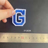 A hand holds a blue and white embroidered letter G patch next to a ruler showing its dimensions.