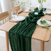 A dark green cheesecloth table runner draped across a light wood table set for an elegant meal.