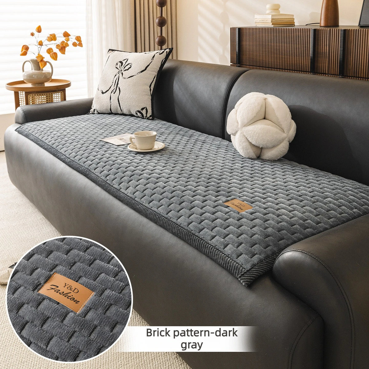 A dark gray brick pattern sofa cover displayed on a leather sofa with a white decorative pillow and a cup.