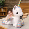 A white unicorn plush toy with a rainbow horn is lying down on a person's lap.