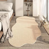 A beige irregular plush area rug placed on a wooden floor next to a bed and a white shelf.