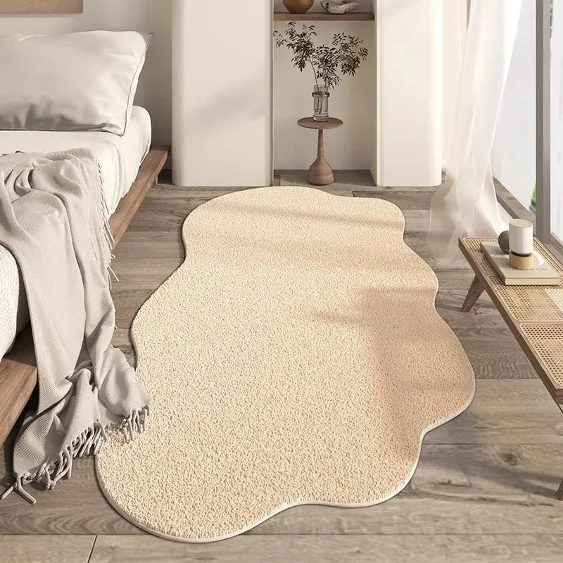 A beige irregular plush area rug placed on a wooden floor next to a bed and a white shelf.