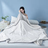 A white Summer Wave Grid Quilt is draped on a bed, with a woman resting her head on the fabric against a light blue wall.