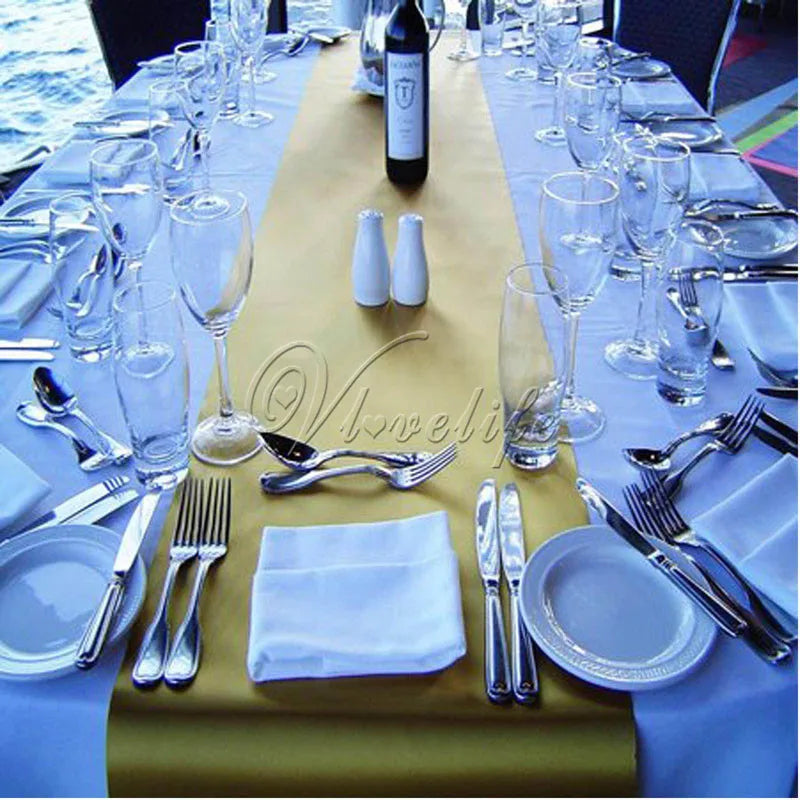 A luxury satin table runner set for an event featuring glassware, cutlery, and a bottle on a long table.