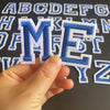 A hand holds an embroidered letter M from a set of alphabet embroidery iron-on patches.