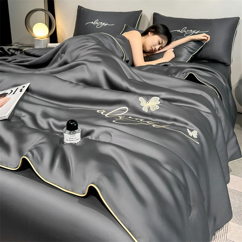 A dark gray ultra-soft ice silk fiber summer quilt draped over a bed with pillows and a person resting.