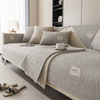 A beige anti-slip sofa cover draped over a light-colored sofa with neutral pillows and a textured throw.