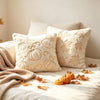 A collection of cream-colored fuzzy maple leaf throw pillows resting on a white bed with a beige blanket and scattered autumn