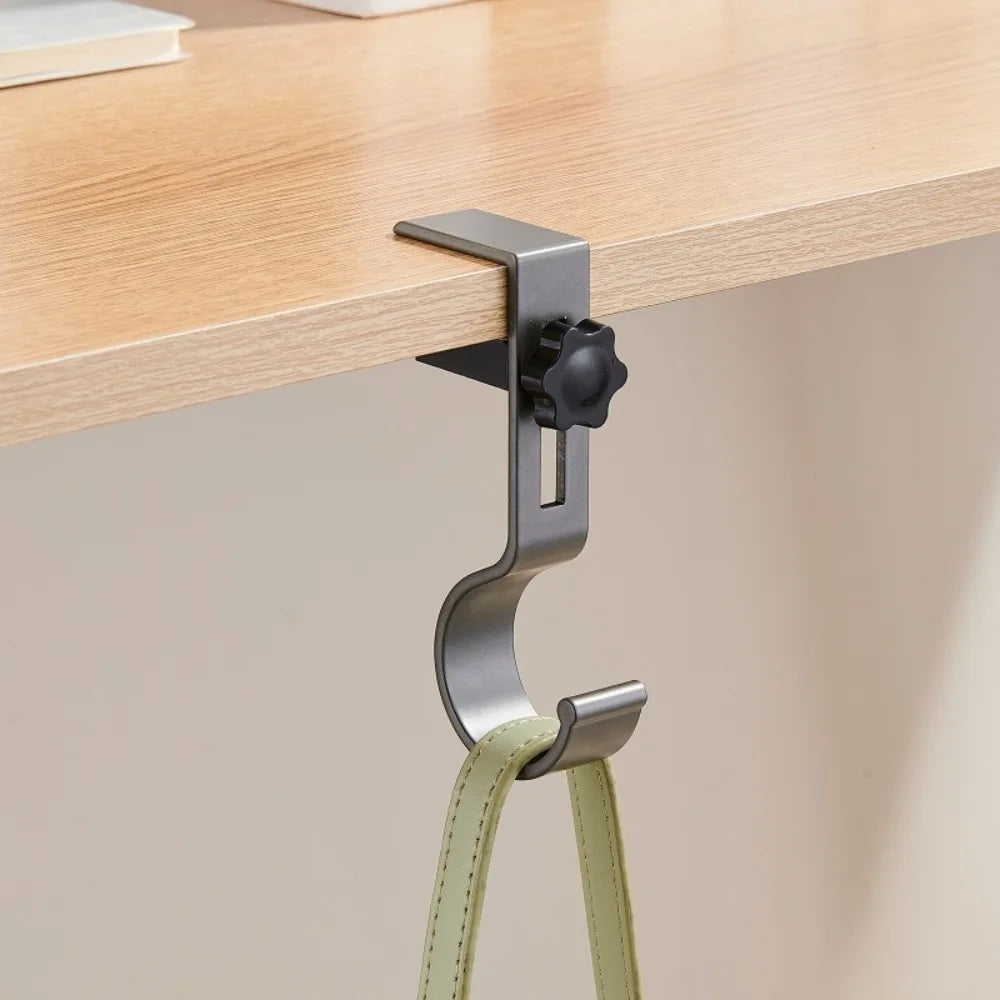 A silver adjustable aluminum desk hook with dual clips is mounted on a wooden shelf.