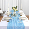 A white table setting featuring a blue gauzy fabric runner, gold candlesticks with white candles, and floral arrangements.