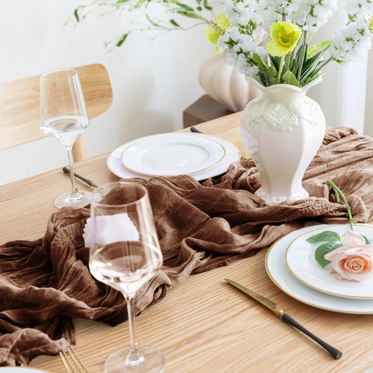 Boho brown cheesecloth table runner draped on a wooden table with glassware and a vase of white flowers.