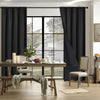 A pair of floor-length black blackout curtains framing a bright window and a wooden dining table.