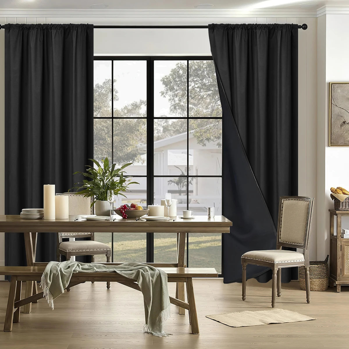 A pair of floor-length black blackout curtains framing a bright window and a wooden dining table.