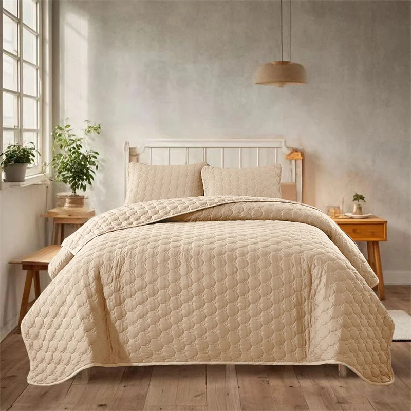 A light beige quilted bedspread set is displayed on a bed with a white headboard in a bright bedroom.