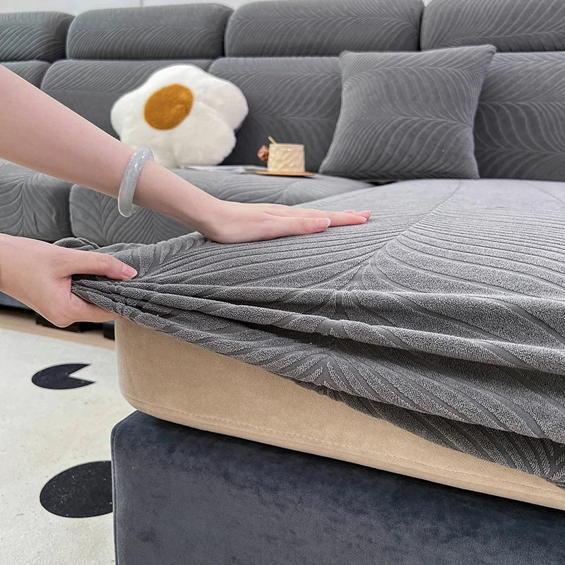 A hand is adjusting a gray jacquard stretch sofa cover on a dark gray sofa, with a white pet-friendly cushion visible.