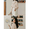 A woman models a cream-colored retro lace long sleeve blouse and white skirt against a light gray wall with framed photos.