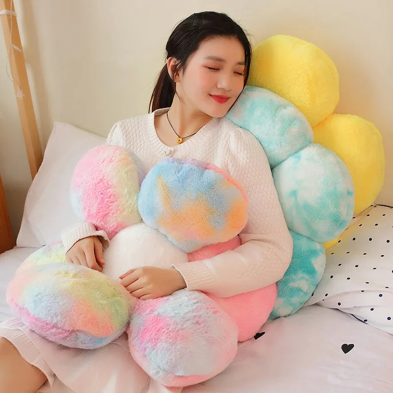 A person is lying down surrounded by various pastel-colored, fluffy plush flowers and cushions.