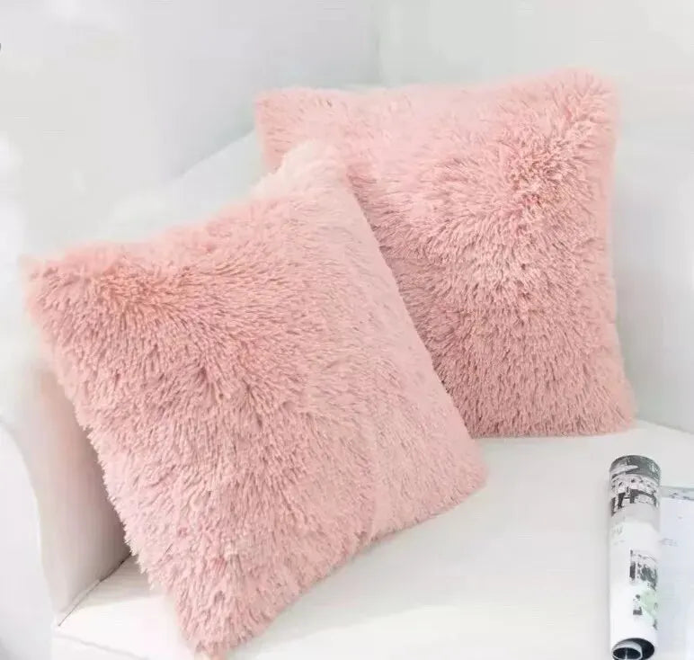 A square, fluffy pink velvet throw pillow resting on a white surface.
