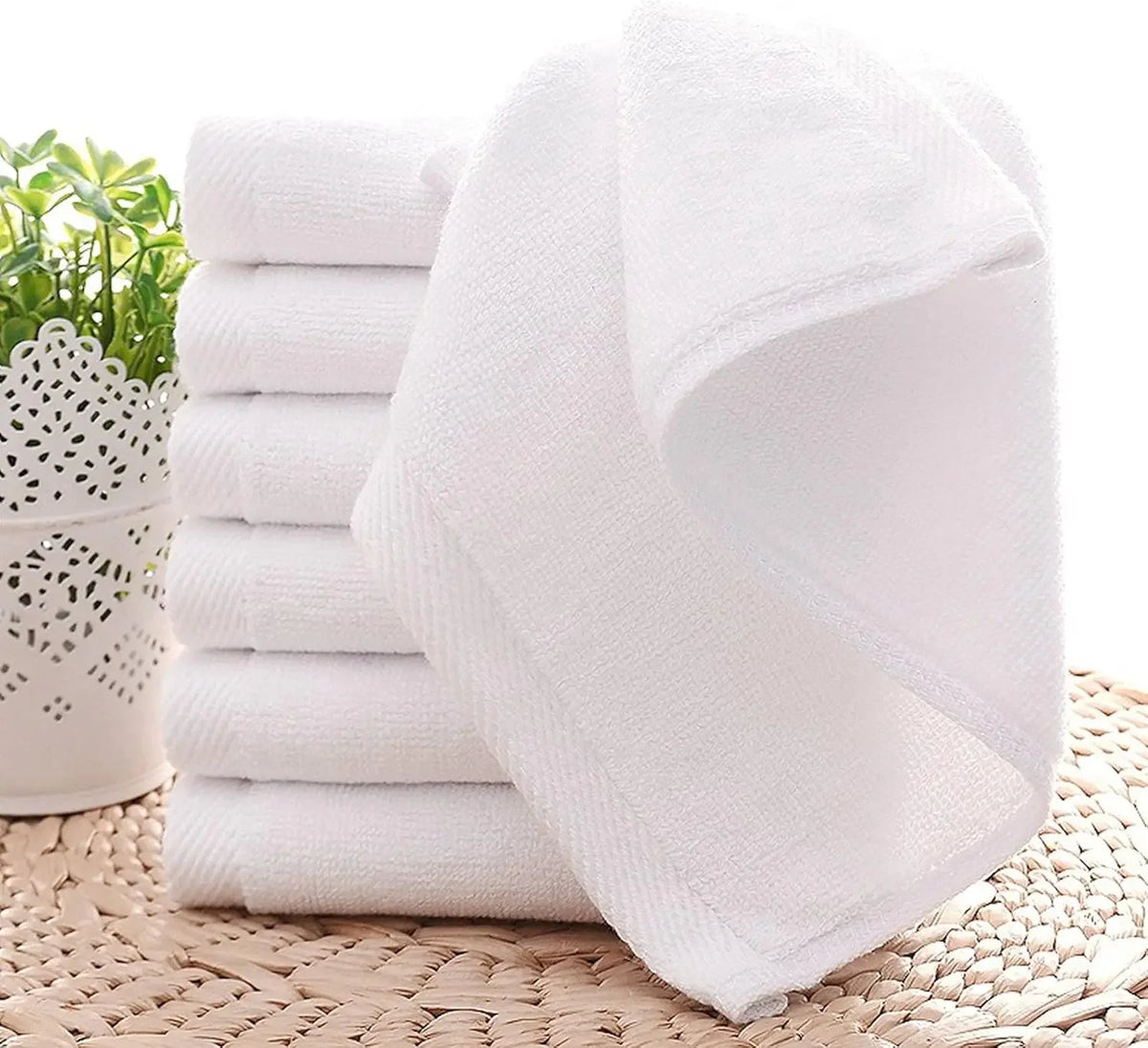 A stack of white cotton face towels is displayed on a woven mat next to some greenery.
