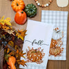 A kitchen towel featuring an autumn pumpkin print with leaves and acorns, set against a wooden background.