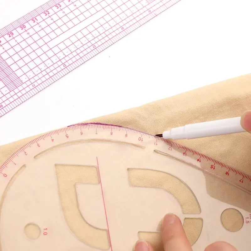 A hand is using a white pen to mark a French curve template on light-colored fabric with a ruler visible nearby.