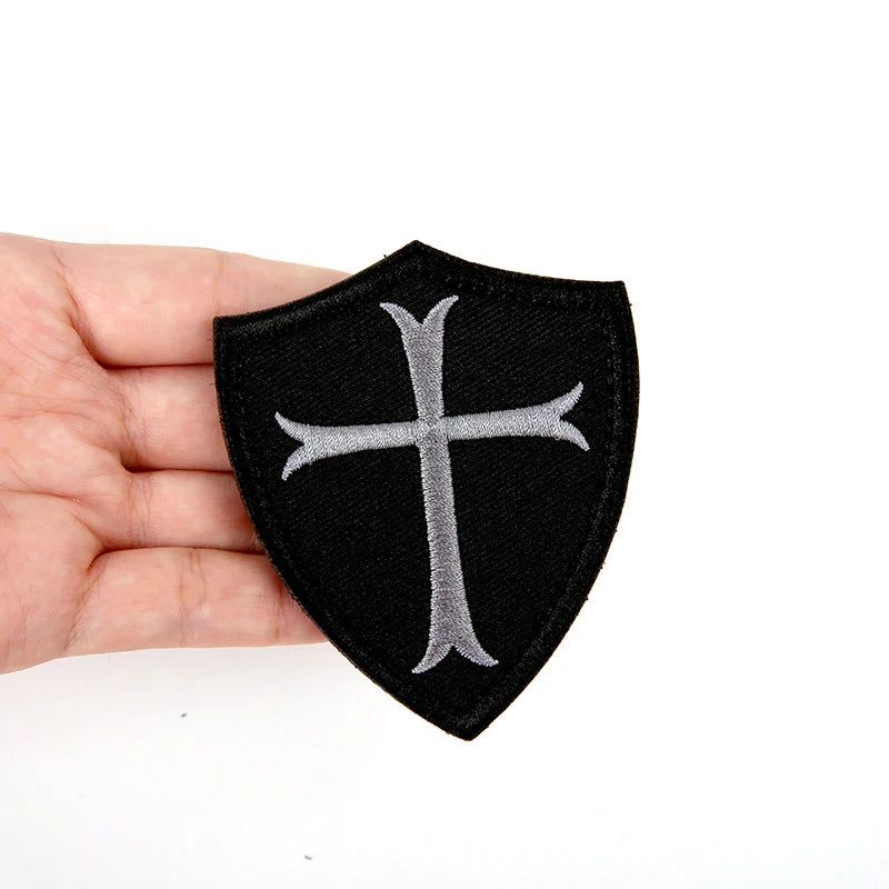 Black embroidered military cross shield patch with hook and loop backing held in a hand.