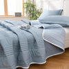Light blue quilted blanket spread over a bed with pillows and a window in the background.