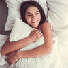 A smiling woman is resting her head on a white bed pillow with a luxurious duvet.