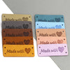 A collection of handmade leather tags in various shades of tan, gray, blue, and pink, each engraved with a heart design.
